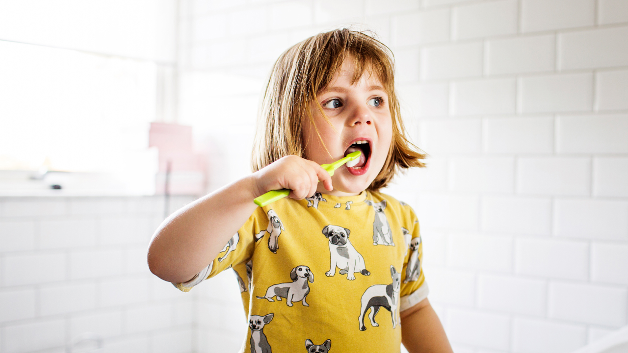 Una bambina bionda si lava i denti in bagno con uno spazzolino verde.